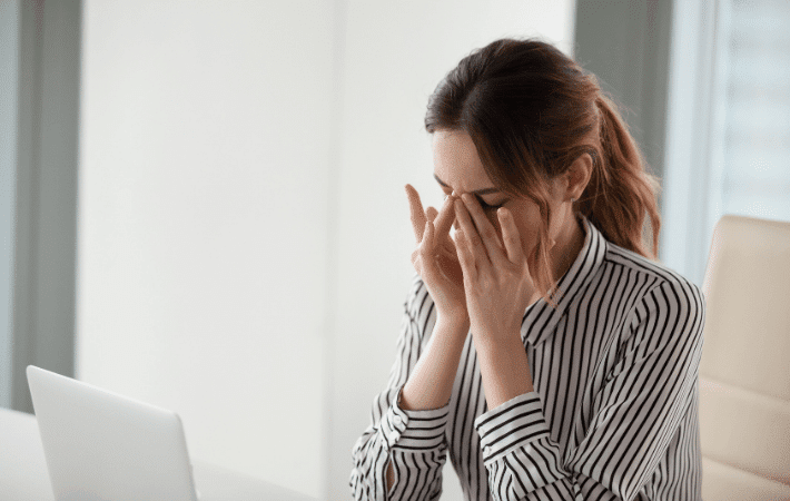 A woman that is working on her laptop and rubbing her strained, dry eyes.