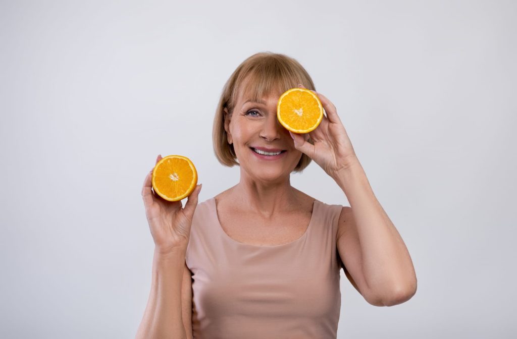 A smiling elderly woman holding orange's in front of her eye's showing that Vitamin C boosts eye health