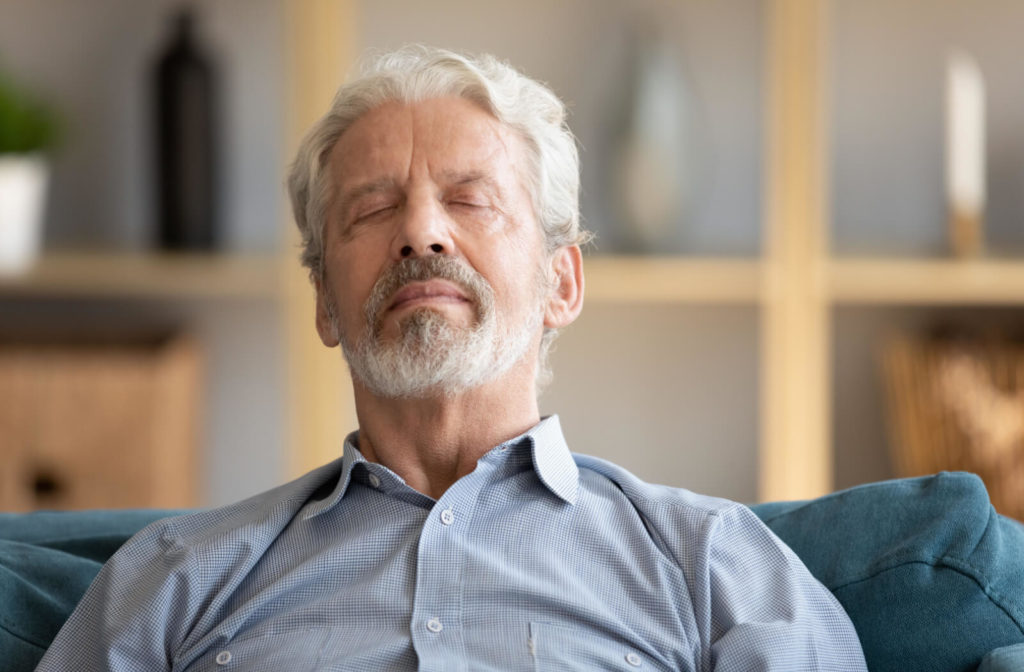 An older man sitting on a sofa with closed eyes, relaxing and taking a break from screen time.