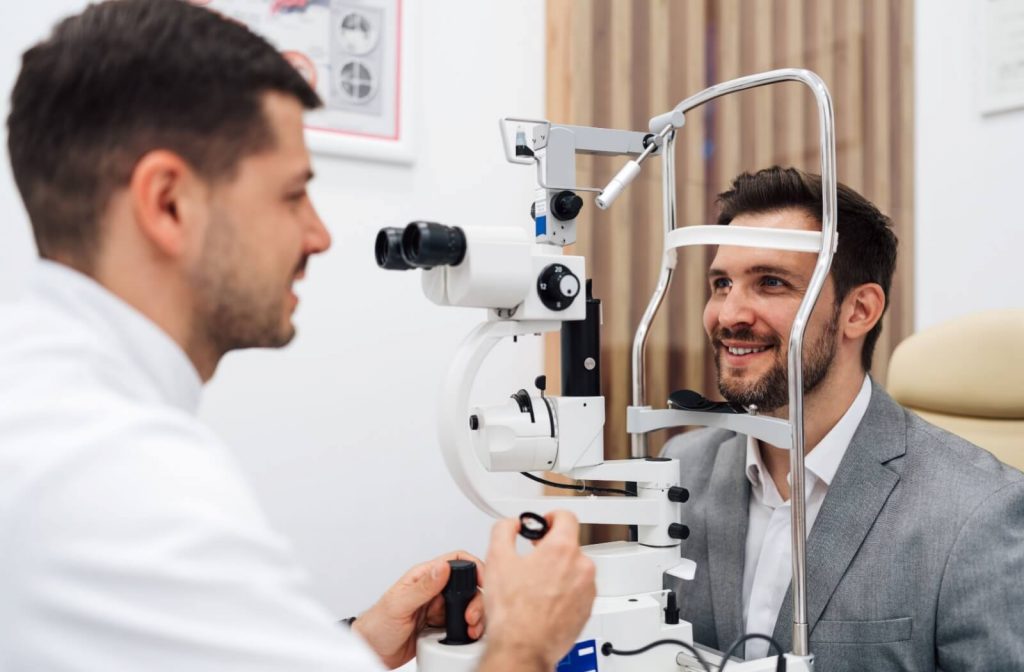 An optometrist performs an eye exam using a slit lamp while a male patient in a gray suit sits and smiles during the test.