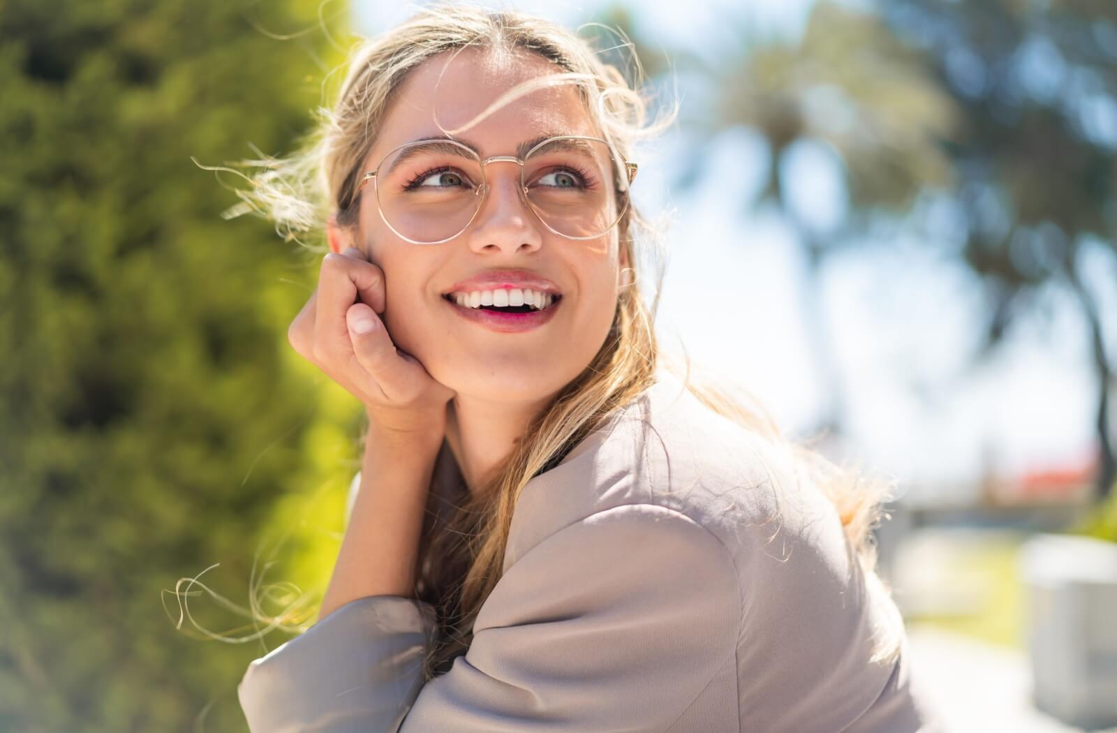 A person is looking away and smiling while resting their chin on their hand and wearing large, round, gold-rimmed glasses outdoors.