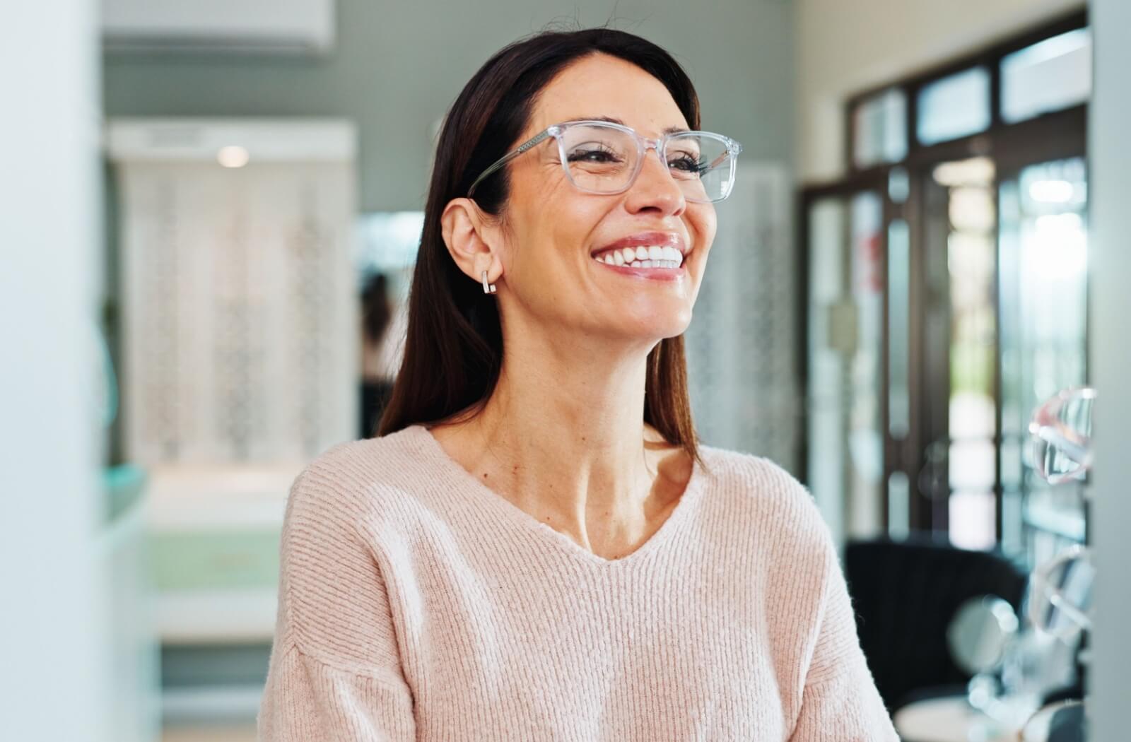 A person smiling while trying on a pair of clear-rimmed glasses in an optical shop.