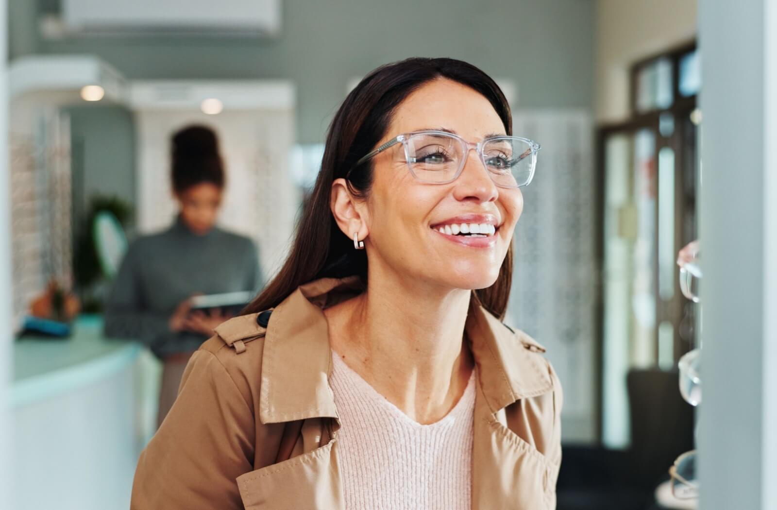 A person wearing a pink sweater and clear-rimmed glasses smiling while looking in a mirror at an optical shop.