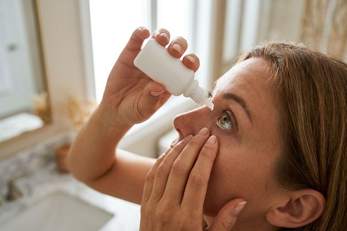 Adult tilting head back and applying eye drops from a white bottle while pulling down lower eyelid, standing in a bright bathroom near a sink